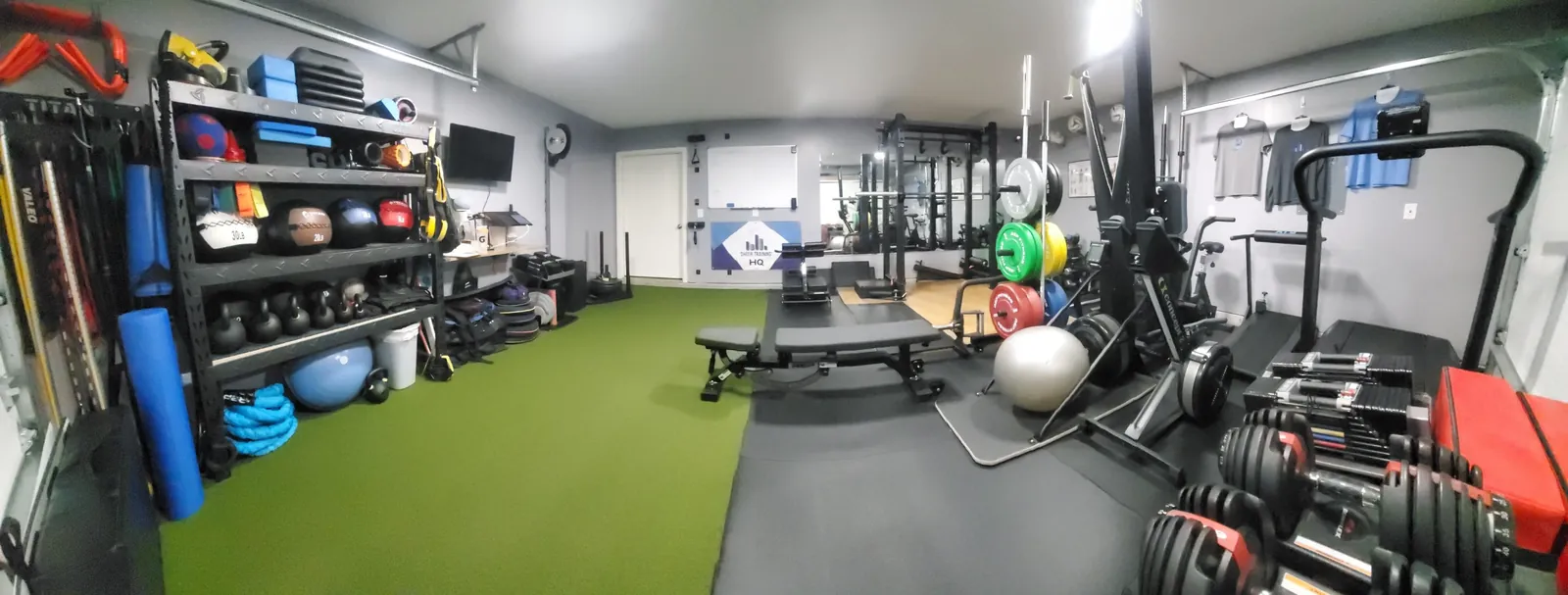Brian’s home gym: a turf lane, squat rack, bumper plates, adjustable dumbbells, and shelving with medicine balls and kettlebells organized for quick use